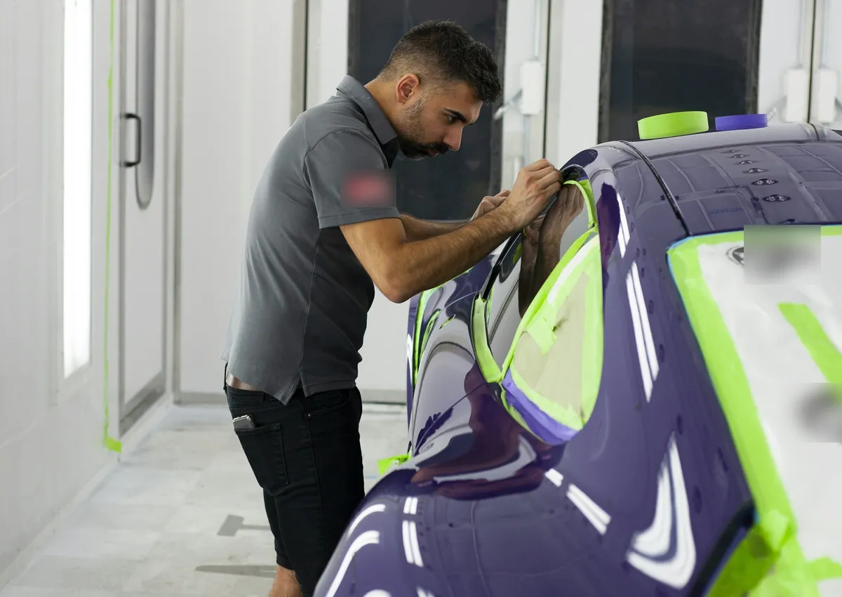 Pintor revisando el enmascarado de un coche morado dentro de una cabina de pintura blanca