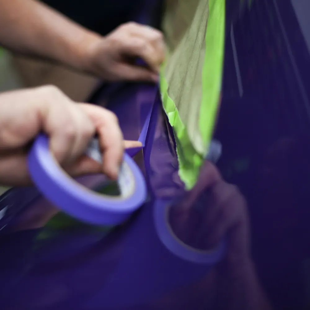 Manos enmascarando con cinta azul una pieza morada antes de pintarla